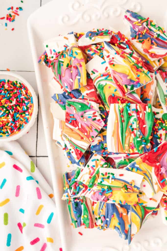 rainbow candy bark on a plate next to a bowl of colorful sprinkles